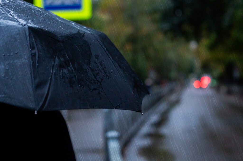 Portrait of a person holding black umbrella outside during the rain. Raindrops and light from city and cars. Rainy weather and forecast concept.